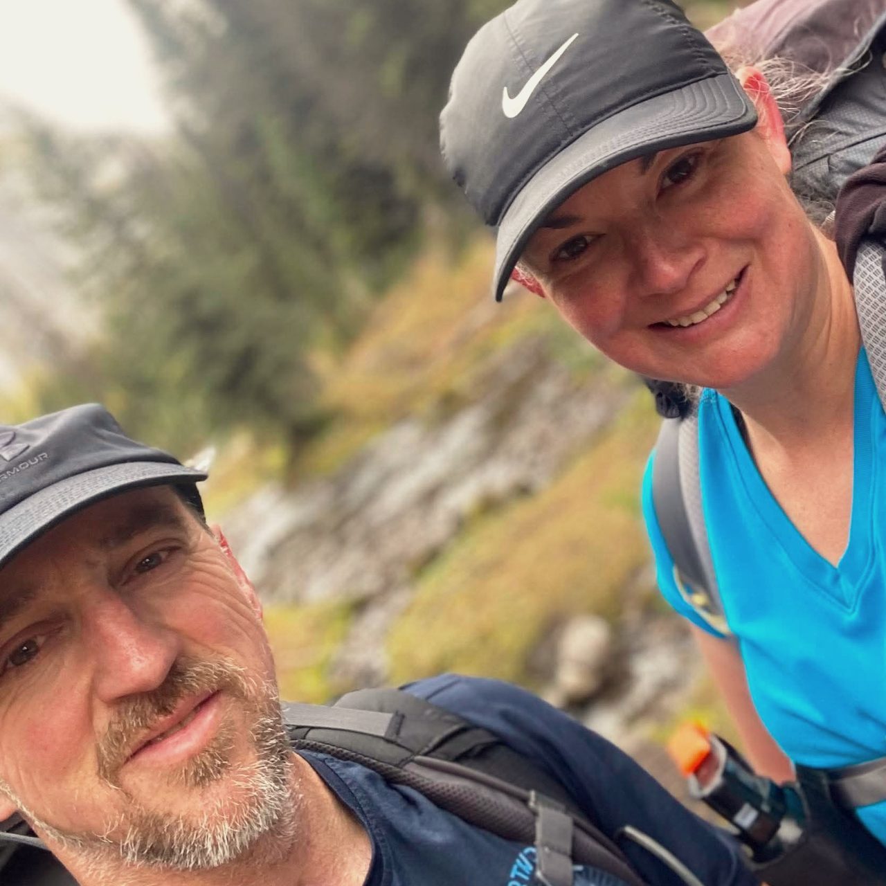 Steve and Emily on a mountain trail, packs on and caps low, smiling into the camera with autumn evergreen slopes behind them